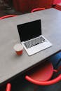 Minimalist workspace with a laptop and coffee cup on a concrete table surrounded by red chairs.